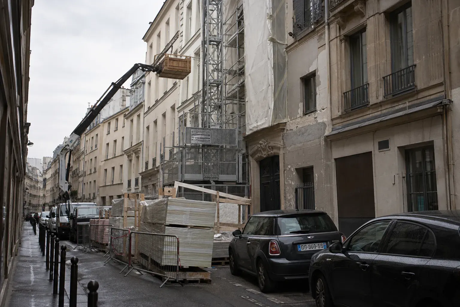Monte-matériaux et petit bras de levage au niveau des toits dans une rue parisienne étroite, palettes de plaques derrière des barrières
