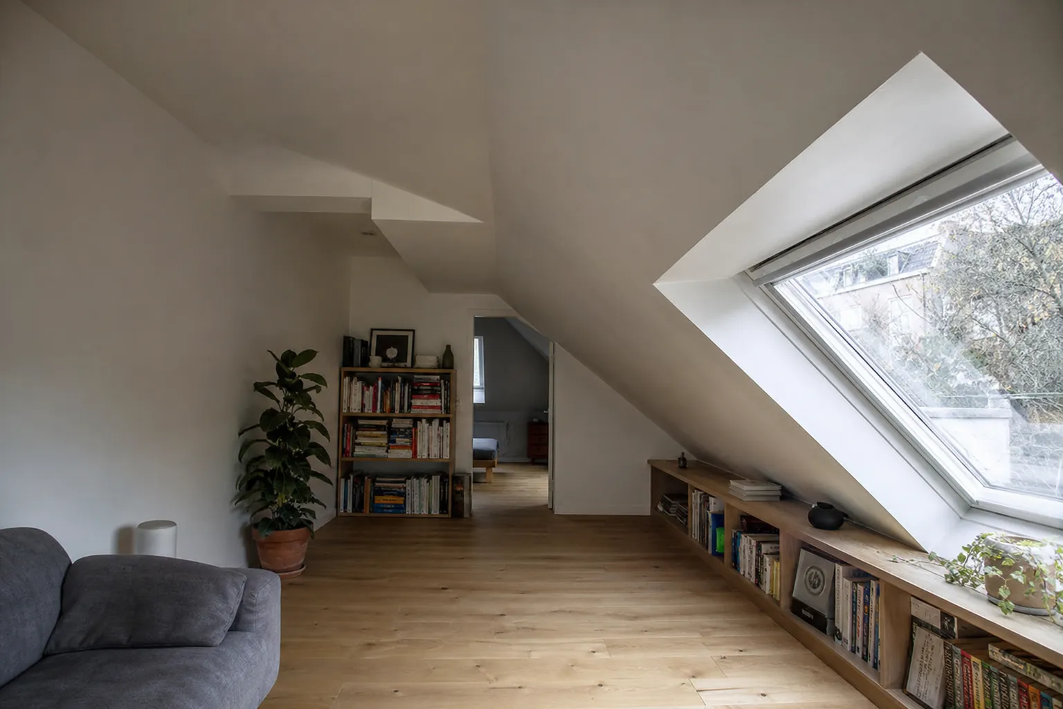 Pièce aménagée sous rampants, murs plâtrés clairs, parquet de chêne et grande fenêtre de toit au-dessus d’une bibliothèque basse