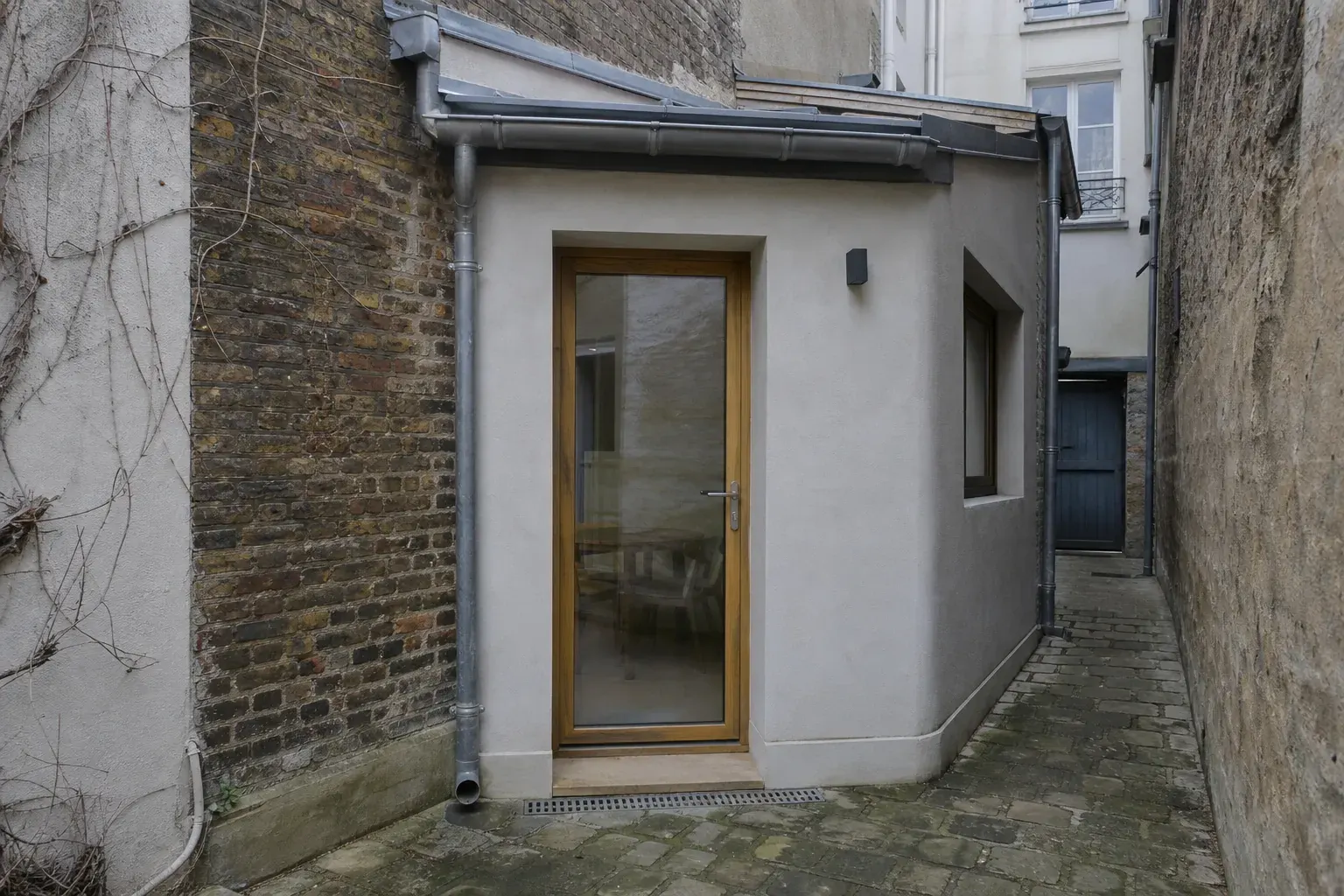 Petite extension de plain-pied ajoutée à l’arrière d’une maison de ville sur une courette pavée, jonction brique et enduit visible